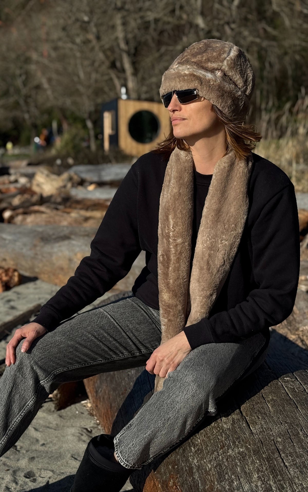 A woman relaxing on a beach log, wearing a Beanie in Golden Bluff Faux Fur with a matching Classic Scarf at golden hour. Made in Seattle, WA, USA by Pandemonium Seattle.
