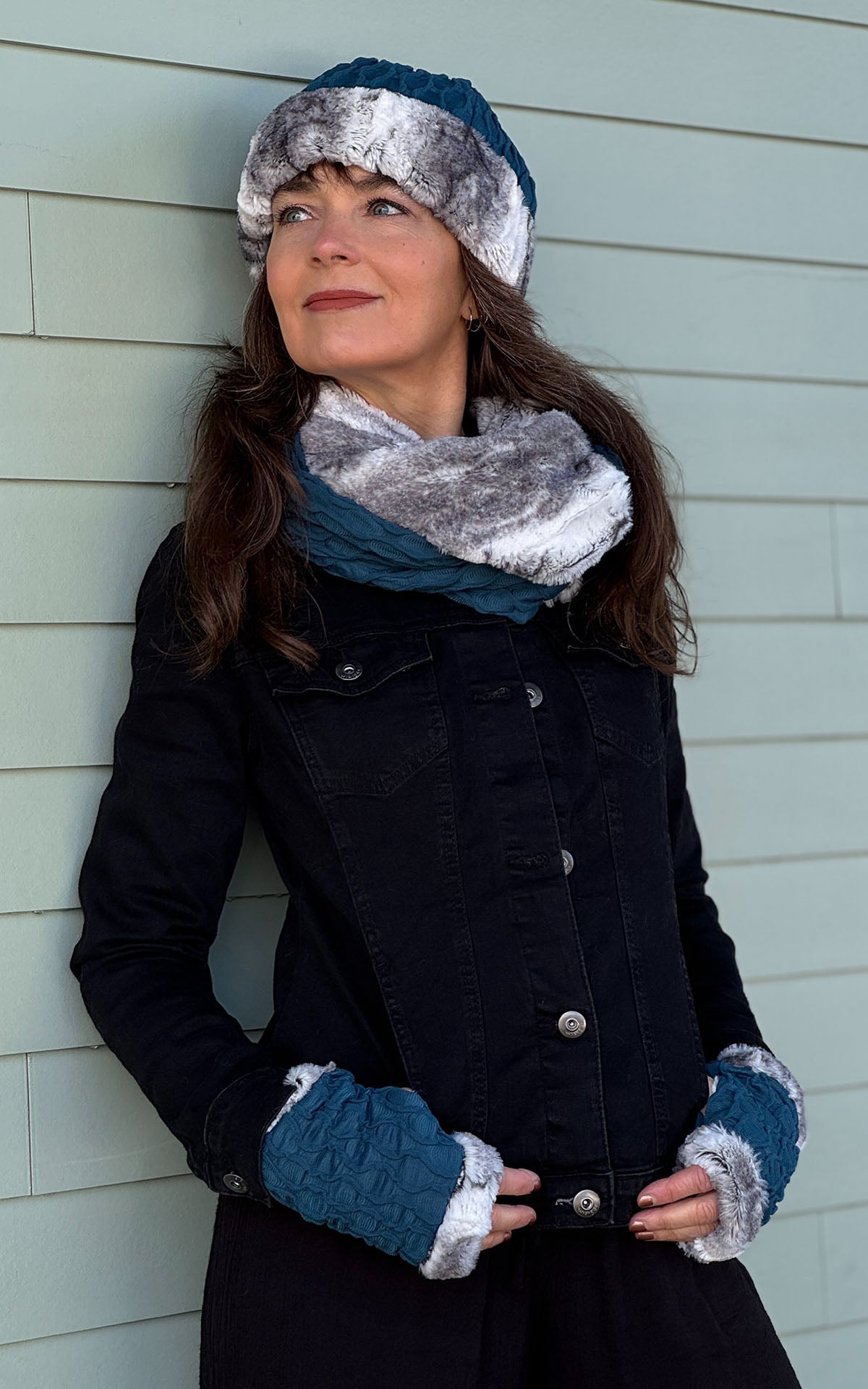 A woman leaning against a wall looking to the left wearing a Two - Tone Infinity Scarf in Cerulean Blue with Smoke Show Faux Fur with matching scarf and fingerless gloves.  Scarf looped around twice on neckline.  Handmade in by Pandemonium Seattle in Seattle WA, USA