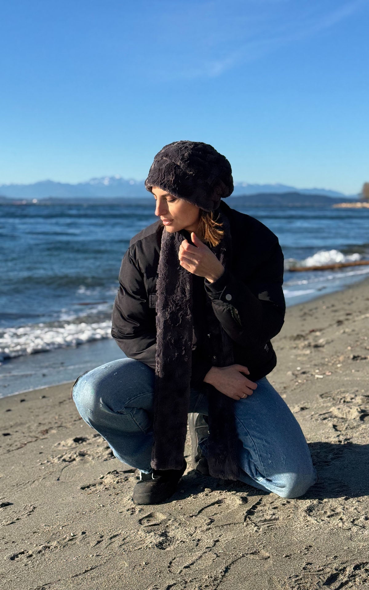 A woman crouching on a sandy beach wearing a Beanie Hat in Smoky Faux Fur, a matching Classic Scarf in Smoky, a black puffer jacket, and blue jeans, with the ocean and mountains in the background. Made in Seattle, WA, USA by Pandemonium Seattle.