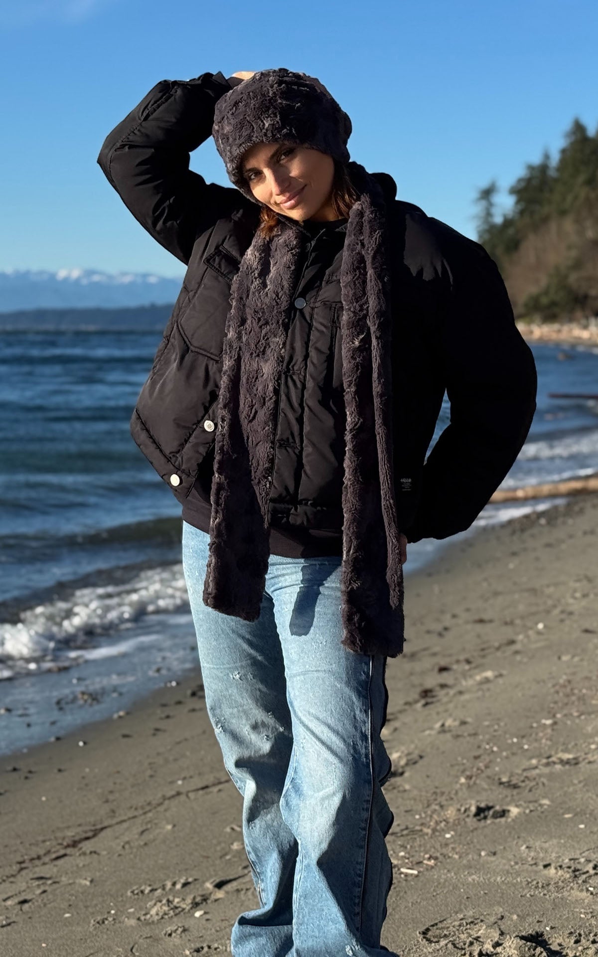 A woman standing on a sandy beach wearing a Beanie Hat in Smoky Faux Fur, a matching Classic Scarf in Smoky, a black puffer jacket, and blue jeans, with the ocean and mountains in the background. Made in Seattle, WA, USA by Pandemonium Seattle.