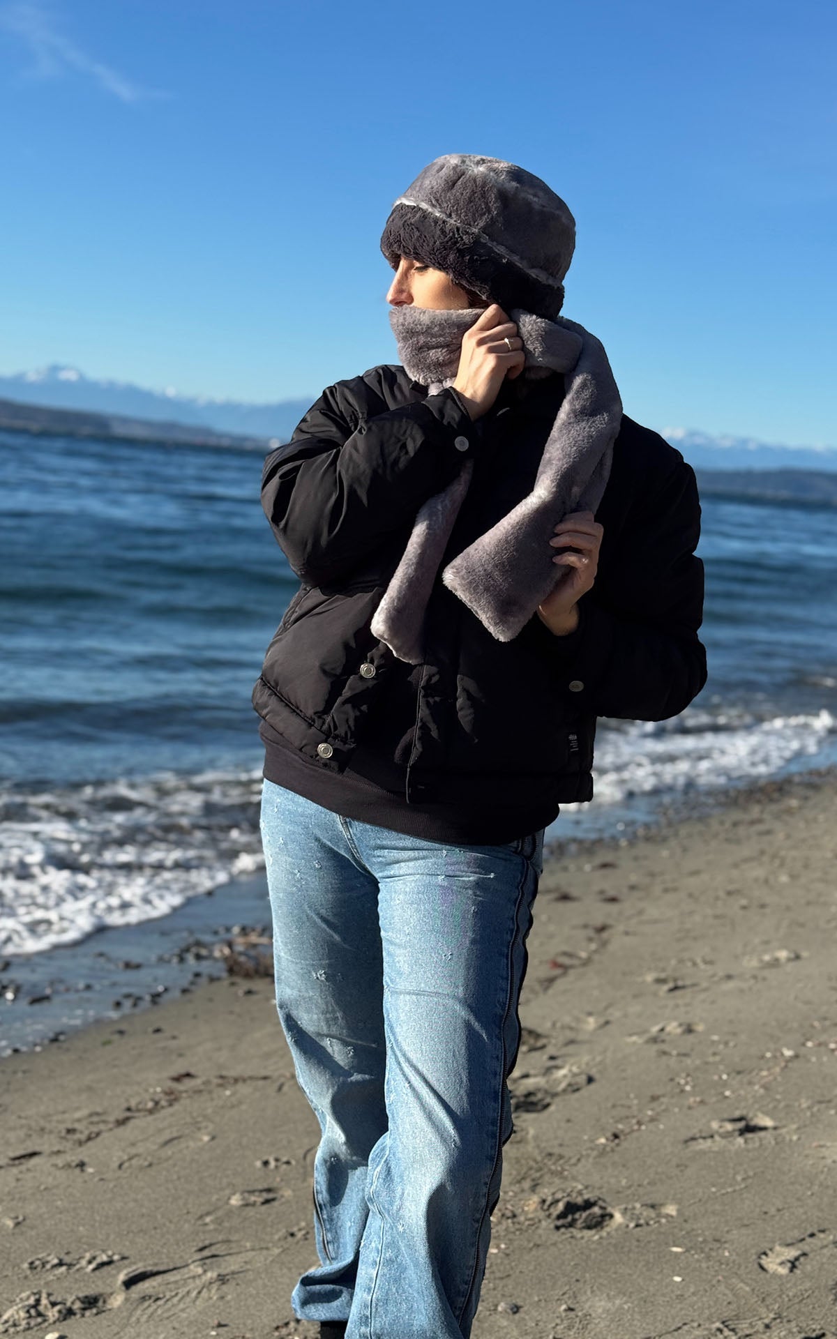 A woman standing on the beach while wearing the Cuffed Pillbox Hat in Olympic Fog with Smoky, with the cuff turned up. Also wearing a matching Olympic Fog Classic Scarf, wrapped around her lower face. Made in Seattle, WA, USA by Pandemonium Seattle.