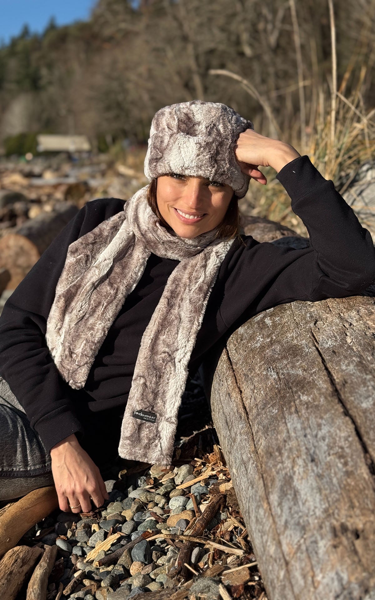 A smiling woman wearing the Cuffed Pillbox in Rusty Rock Faux Fur with a matching Classic Scarf, while resting against a beach log. Made in Seattle, WA, USA by Pandemonium Seattle.