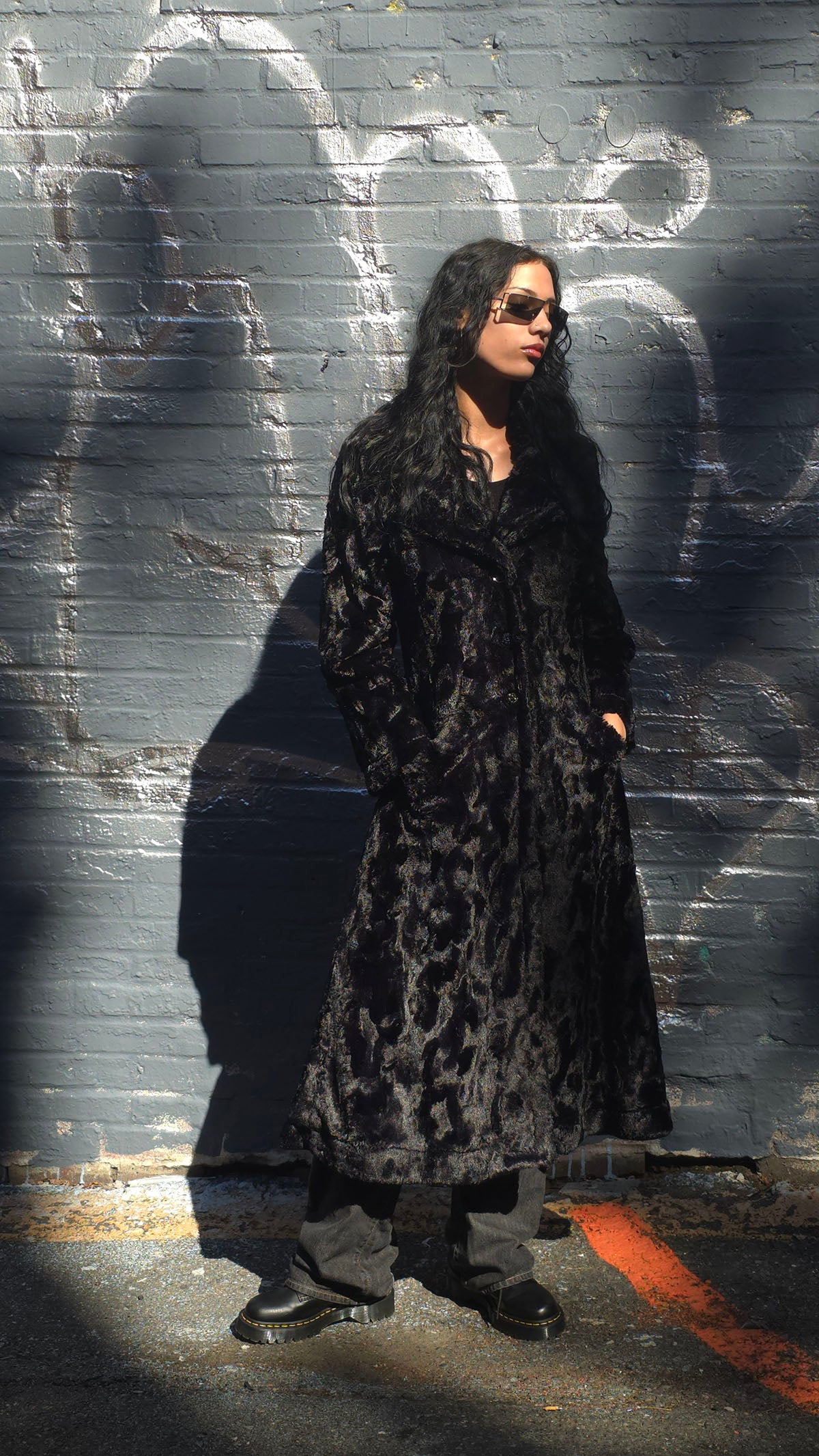 Woman wearing the Hayworth Coat in Cuddly Black with hands in pockets, while standing near a brick wall. Made in Seattle, WA, USA by Pandemonium Seattle.