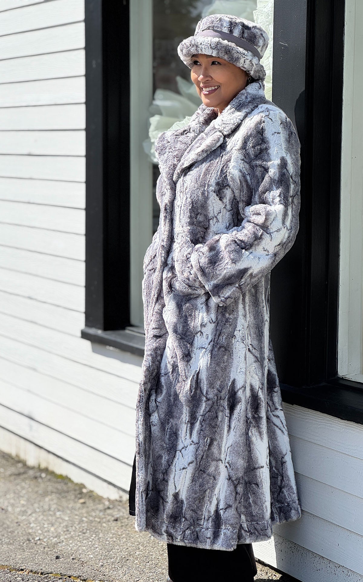 Side view of a woman leaning against a  building with wearing  the Hayworth Coat in Smoke Show Faux Fur with a matching Faux Fur Molly Bucket Style Hat in Smoke Show. Handmade in Seattle WA, USA by Pandemonium Seattle