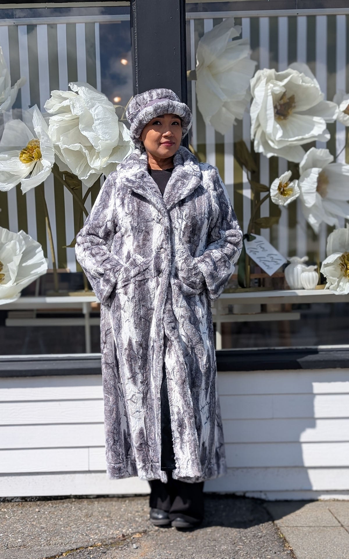 Woman leaning against a window with white floral display featuring the Hayworth Coat in Smoke Show Faux Fur with a matching Molly hat. Handmade in Seattle WA, USA by Pandemonium Seattle