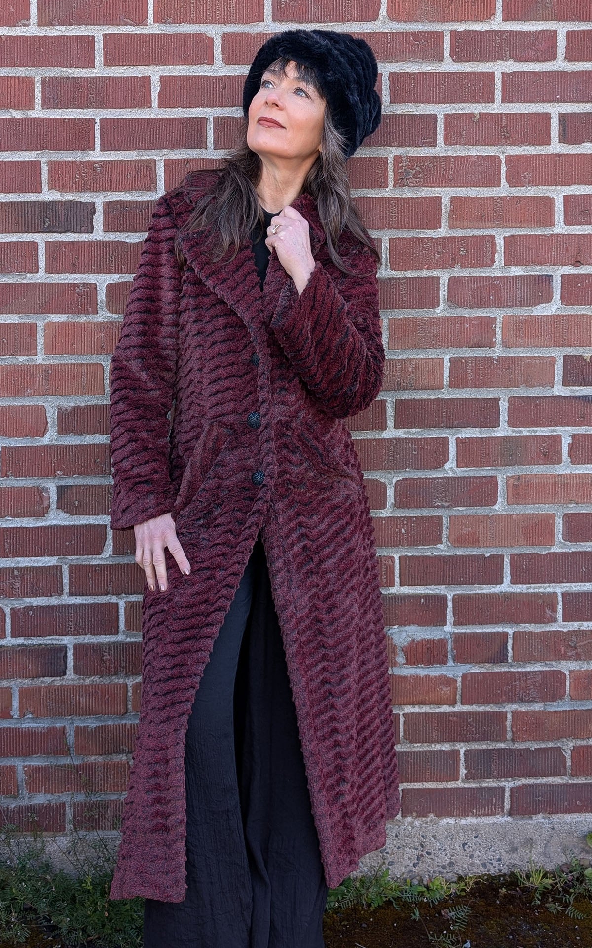 A woman looking up, while wearing the Hayworth Coat in Desert Sand in Crimson Faux Fur and a Beanie hat in Onyx Faux Fur. Handmade in Seattle WA, USA by Pandemonium Seattle