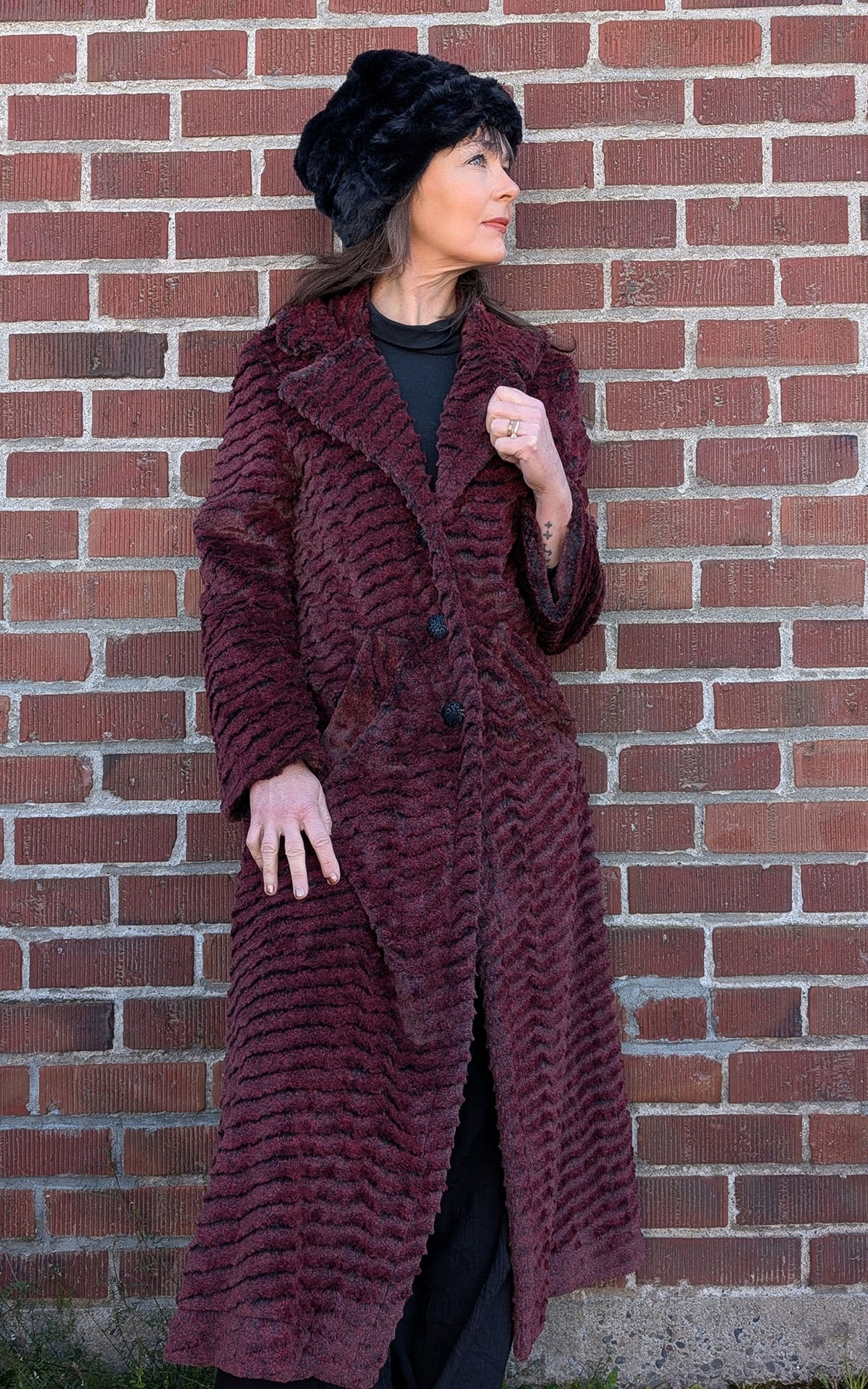 A woman leaning against a brick wall wearing the Hayworth Coat in Desert Sand in Crimson Faux Fur. Handmade in Seattle WA, USA by Pandemonium Seattle