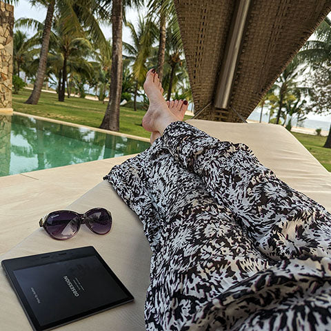 woman's legs in silk pants lounging by poolside with glasses and tablet