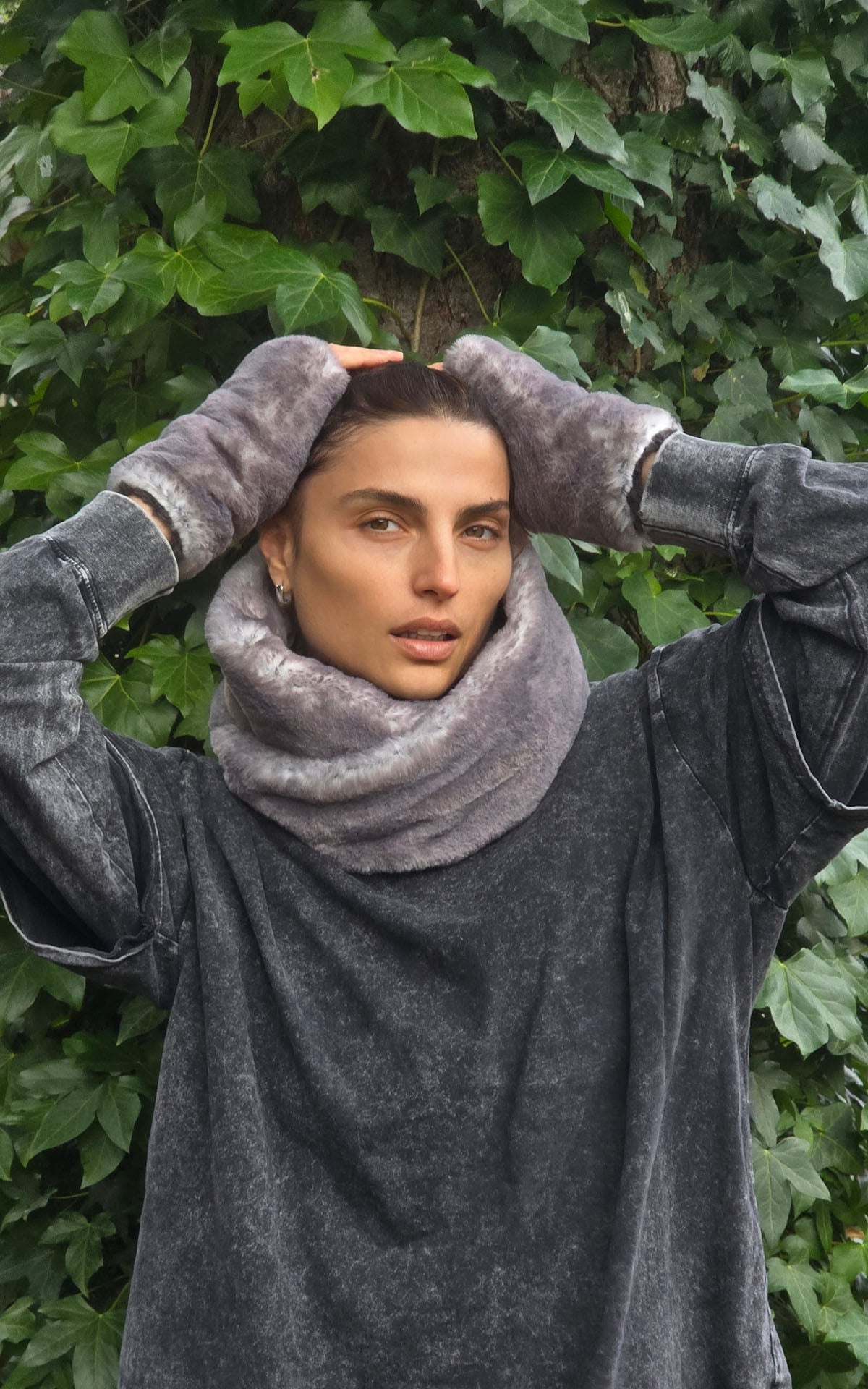 A woman wearing a plush, fuzzy gray cowl neck warmer in Olympic Fog Faux Fur and matching fingerless gloves, standing in front of a background of green ivy. Made in Seattle, WA, USA by Pandemonium Seattle.