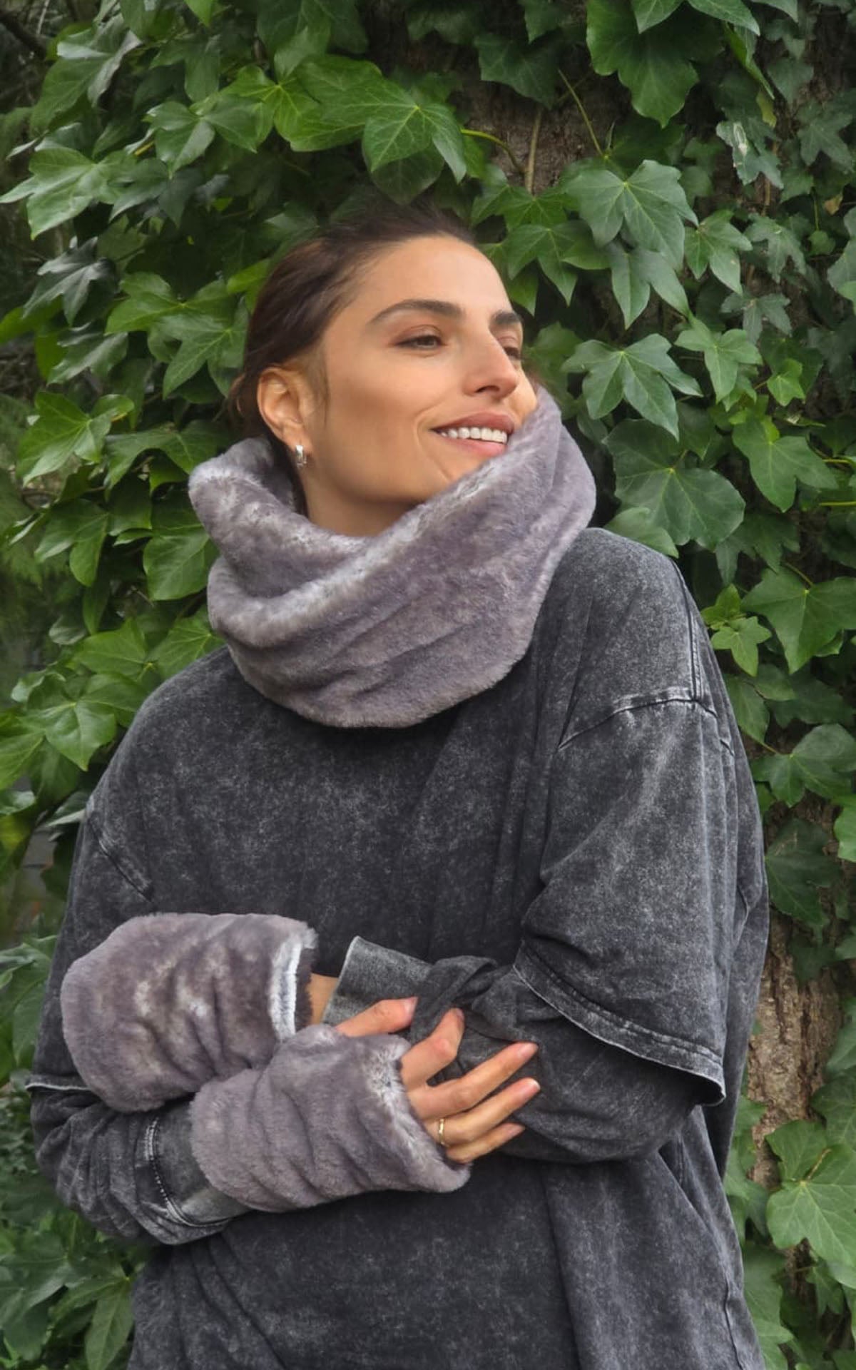 A smiling woman wearing a plush, fuzzy gray cowl neck warmer in Olympic Fog Faux Fur and matching fingerless gloves, standing in front of a background of green ivy. Made in Seattle, WA, USA by Pandemonium Seattle.