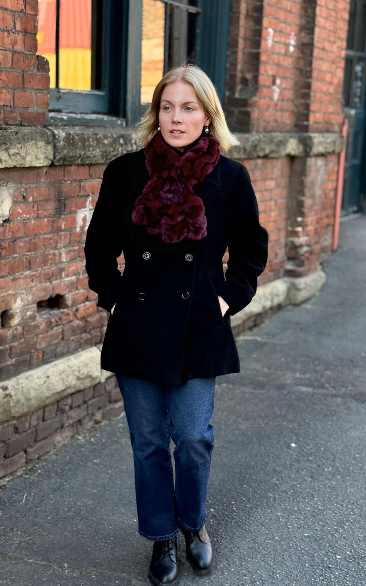 A woman standing outdoors on a sidewalk next to a brick building, wearing a black double-breasted coat, blue jeans, and the Royal Opulence Pull-Thru Scarf in Cherry Bomb. Made in Seattle, WA, USA by Pandemonium Seattle.