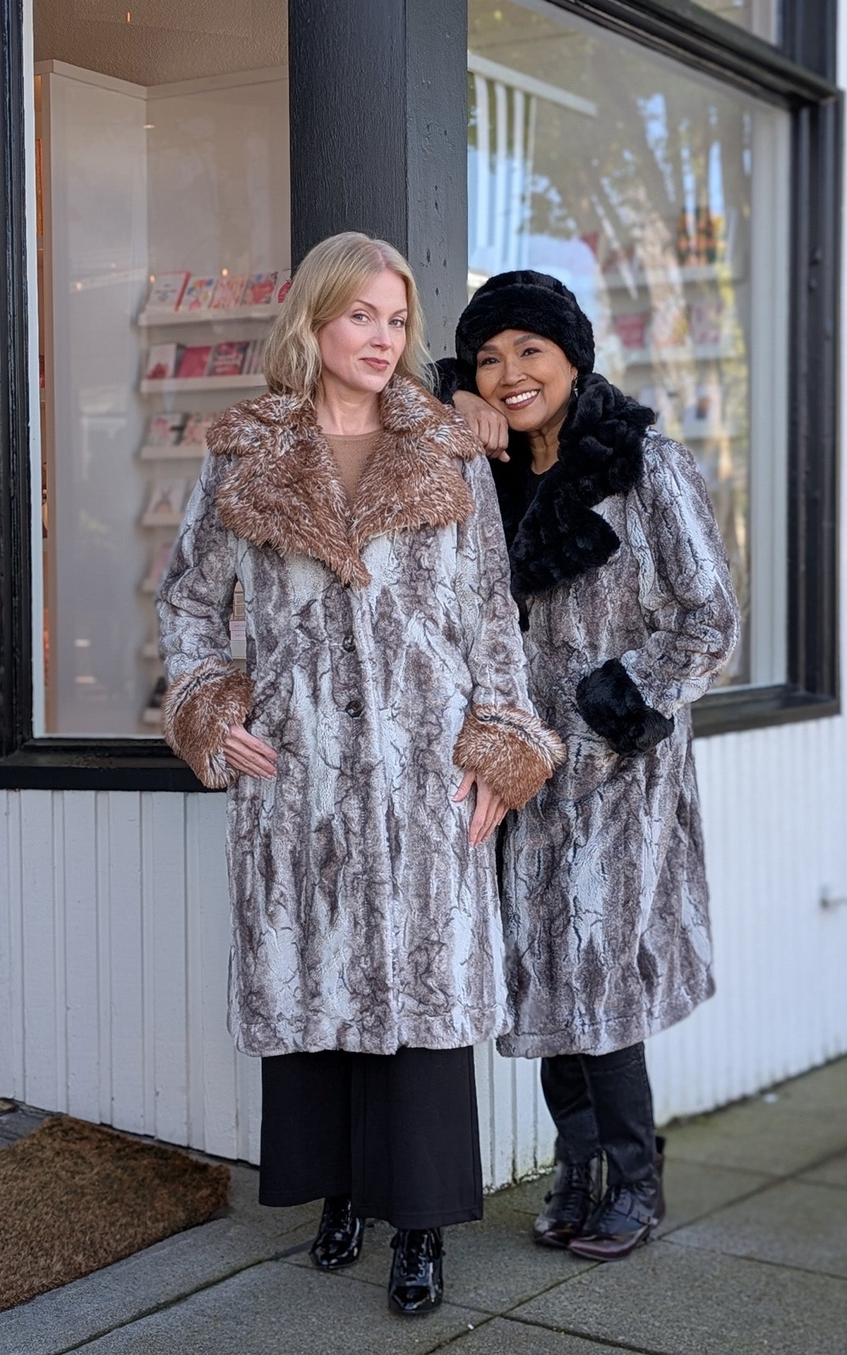 Two women modeling coats, on the right the woman resting on the other women's shoulder. She features the Roxy Coat - Rocksteady Faux Fur with Onyx collar and cuffs, and the other is in the Rusty Rock  Faux Fur with Red Fox collar and cuffs.   Handmade by Pandemonium Seattle, USA should. 