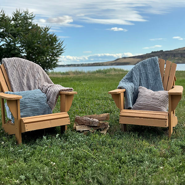 Two luxury faux fur throws in 'Honey' and 'Blue Mist' colors draped over outdoor wooden chairs with a scenic lake in the background.