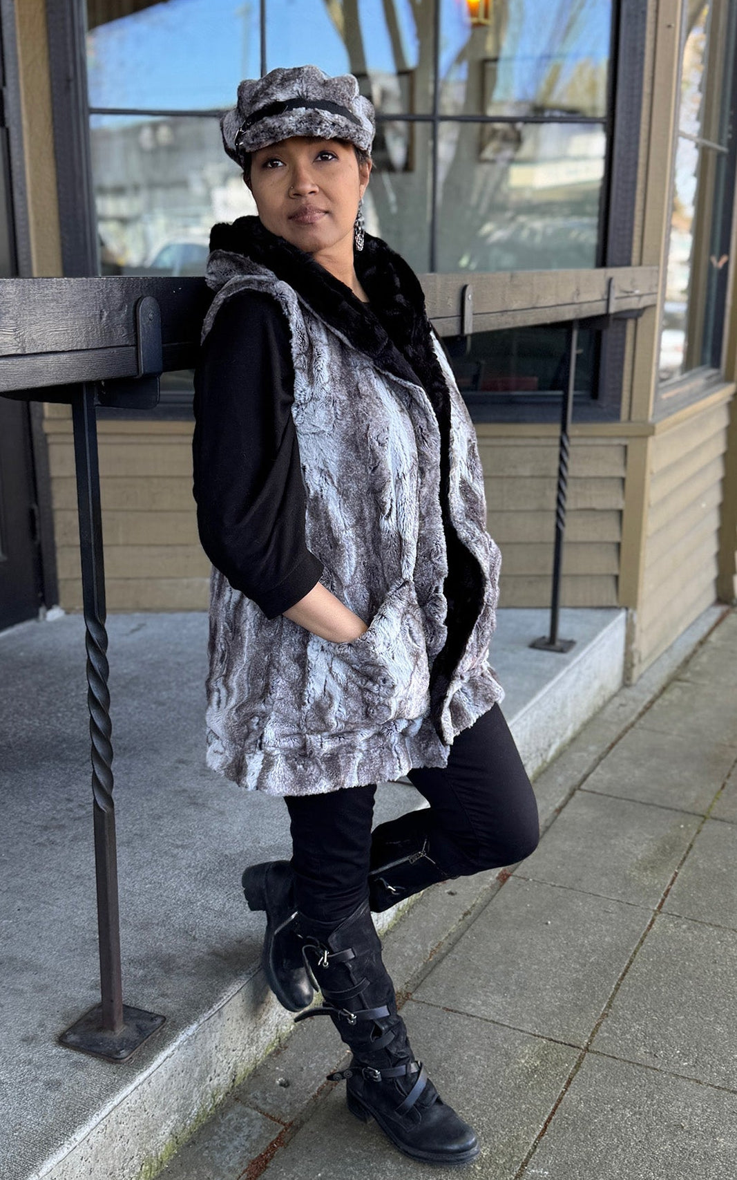 A woman leaning on a railing wearing the Oversized Hooded Vest in Rock Steady Faux Fur with hands in pocket.  She features a matching Valerie cap Styled hat.  Handmade in Seattle WA, USA by Pandemonium Millinery