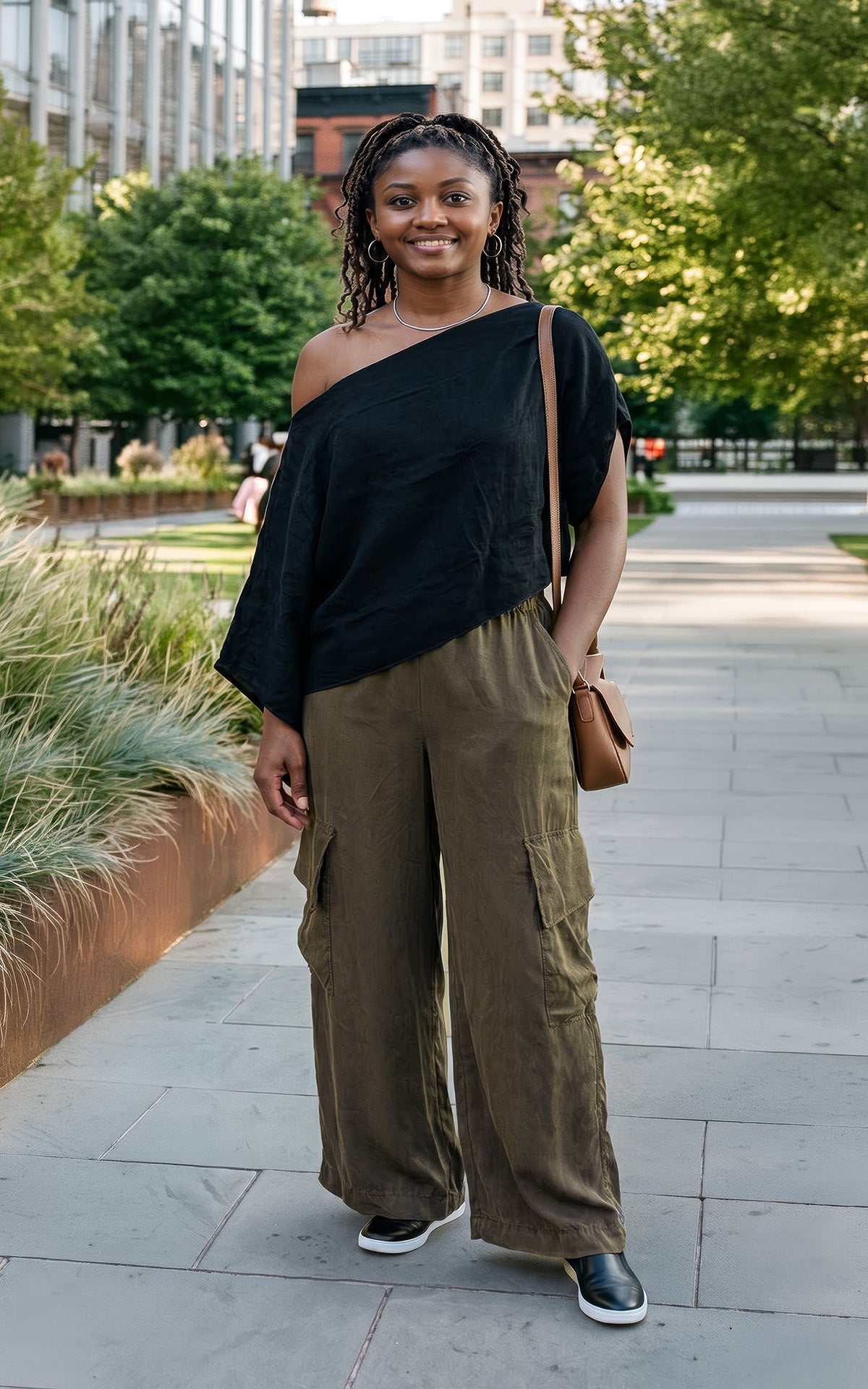 A woman stands on a paved outdoor walkway wearing a black off-the-shoulder top and olive green wide-leg cargo pants, with a small brown shoulder bag and black-and-white sneakers. Made in Seattle, WA, USA by Pandemonium Seattle.