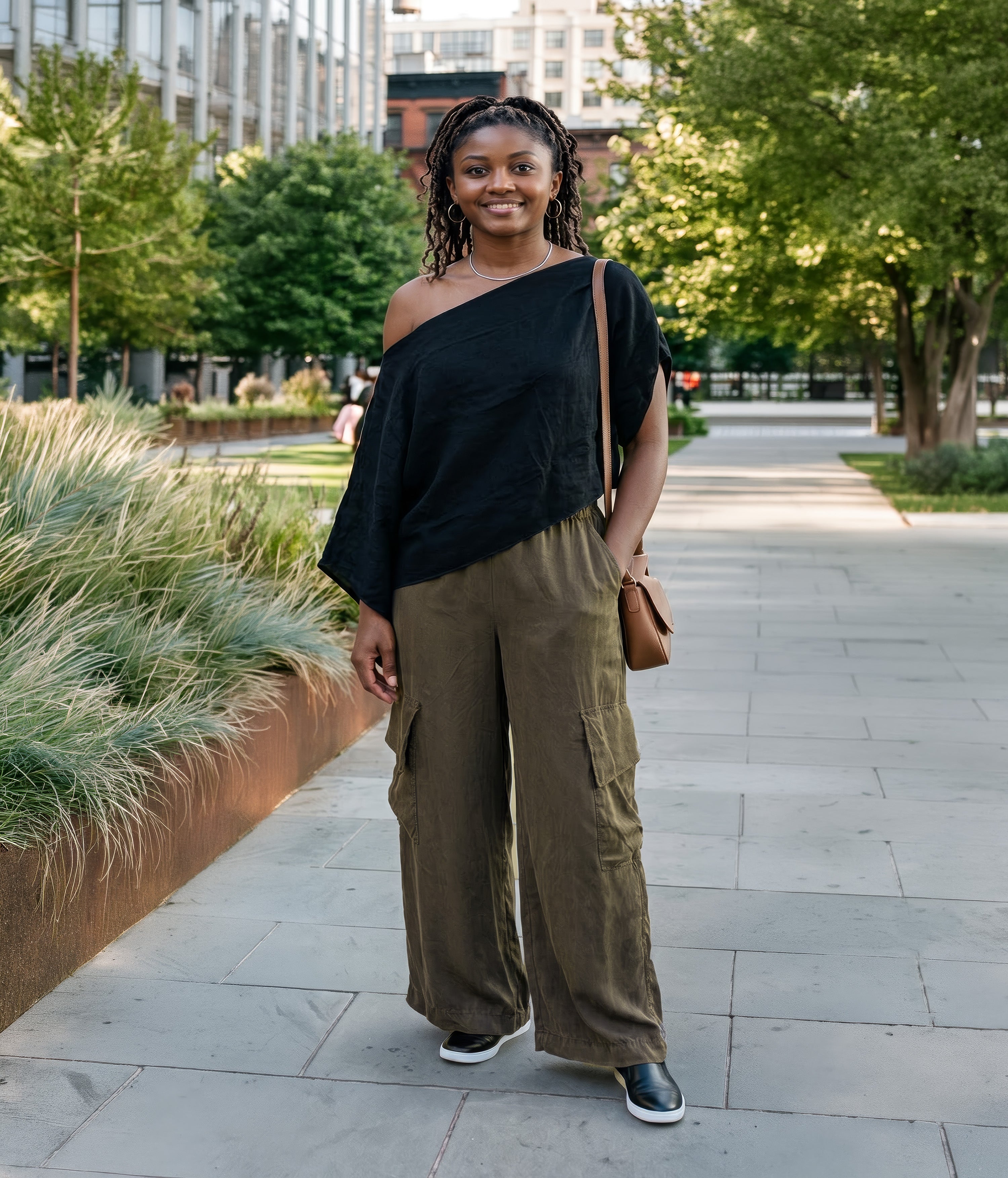 A woman stands on a paved outdoor walkway wearing a black off-the-shoulder top and olive green wide-leg cargo pants, with a small brown shoulder bag and black-and-white sneakers. Made in Seattle, WA, USA by Pandemonium Seattle.