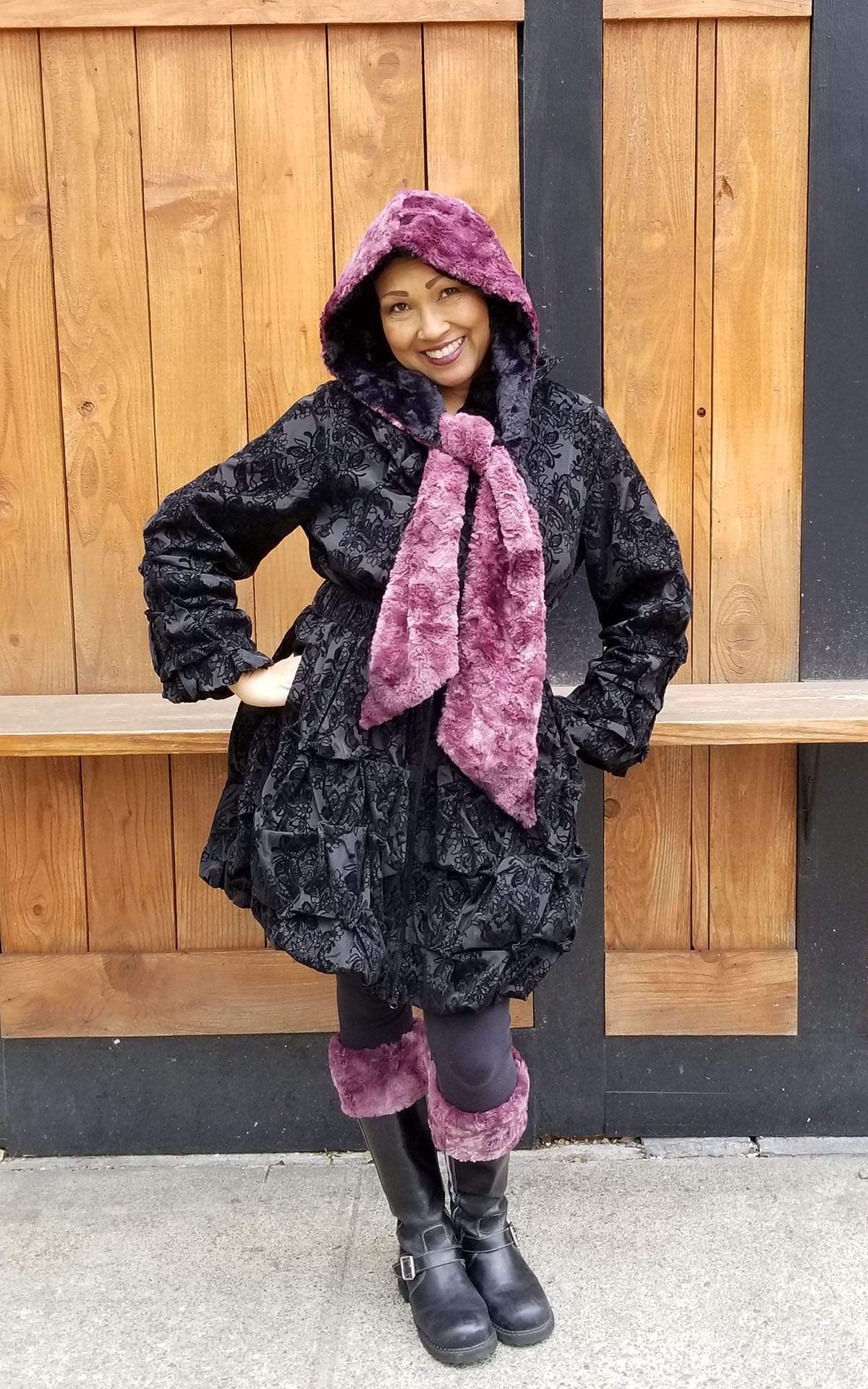 Model standing in front of wood fence wearing women’s Boot toppers and matching  Two-Tone Hooded Scarf | Highland in Thistle, Mauve reversing to Cuddly Fur in Sand, Ivory Faux Fur | Handmade in Seattle WA | Pandemonium Millinery