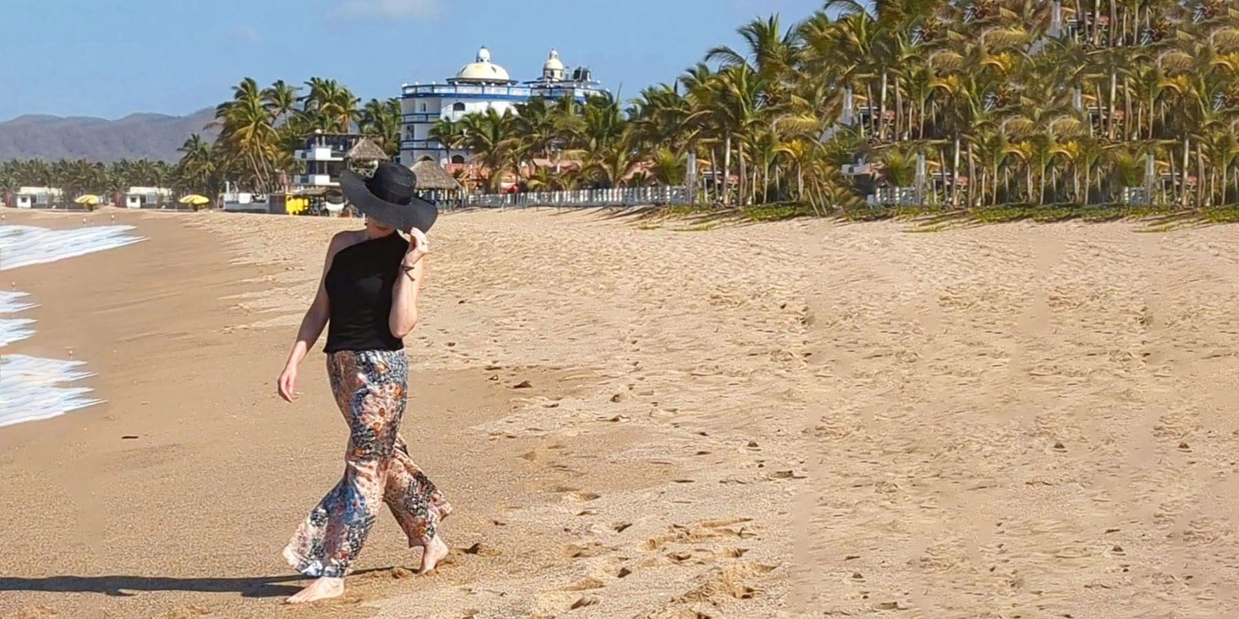 model walking on beach in floral silk palazzo pants with sun hat handmade in the USA by Leigh Young Collection a division of Pandemonium Seattle