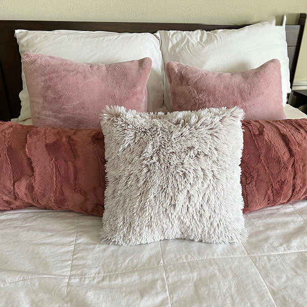 A variety of handmade faux fur pillow shams in neutral and soft pink tones styled on a white bed, showcasing different textures and sizes