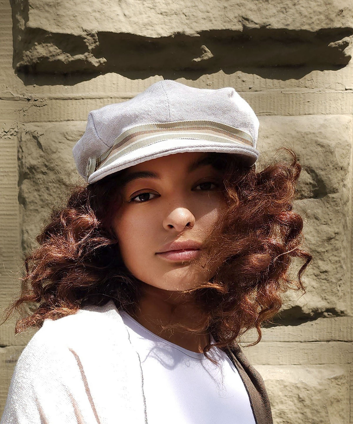 Model against a brick wall wearing Valerie Cap Style Hat in Linen in Bone | Multi-Stripe Band | Handmade by Pandemonium Millinery | Seattle WA