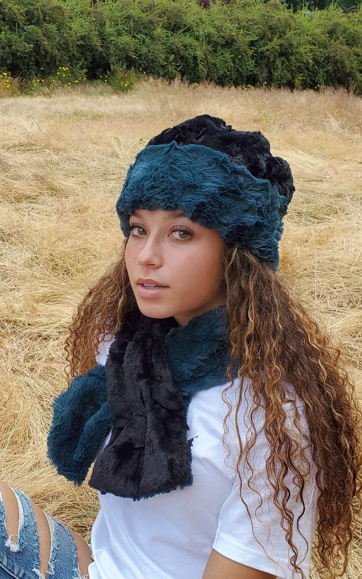 Model sitting in a field wearing a reversible Beanie and a matching  Women’s Pull Through Scarf shown in reverse Peacock Pond Faux Fur, Blue / Teal with Cuddly Faux Fur in Black | Handmade in Seattle WA | Pandemonium Millinery