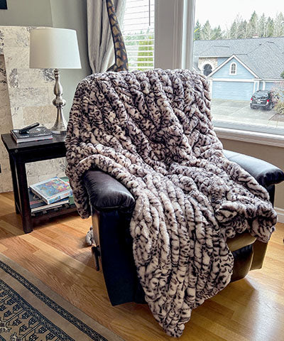 Draped over a chair is a  Luxury Faux Fur Throw in Snow Leopard From Royal Opulence Collection made by Pandemonium Seattle